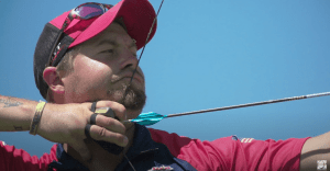 An Olympic archer in a red shirt is pulls back the string of their bow.